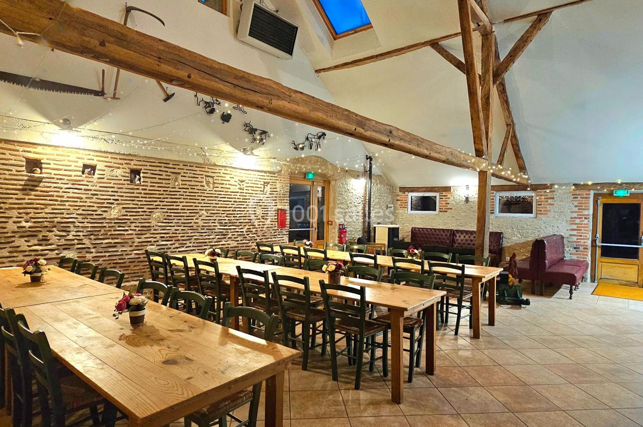 Salle lumineuse avec murs en briques, poutres apparentes, tables en bois et chaises alignées pour un événement.