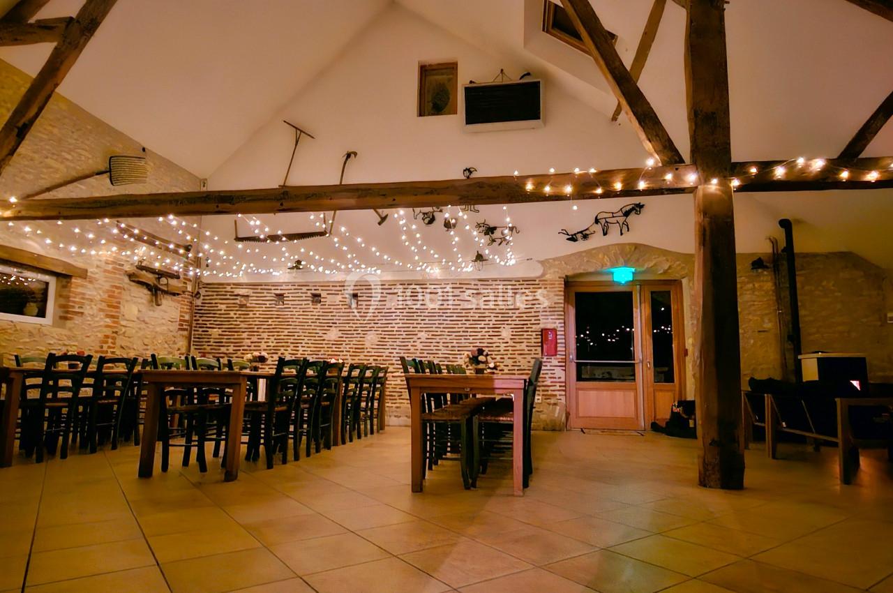 Salle lumineuse avec murs en briques, poutres apparentes, guirlandes suspendues et tables en bois alignées.