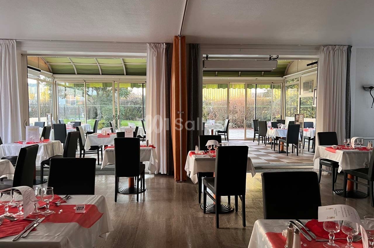 Salle de restaurant lumineuse avec tables dressées, nappes blanches et chaises noires, donnant sur une terrasse vitrée.