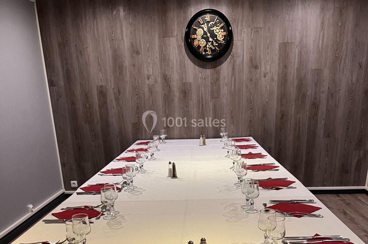 Salle à manger avec une grande table dressée, nappée de blanc, couverts alignés et horloge murale en arrière-plan.