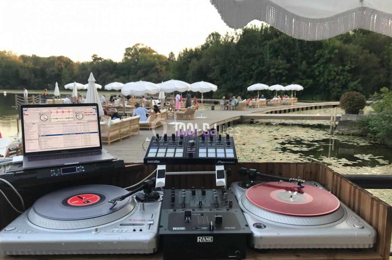 Poste de DJ en plein air avec platines et ordinateur, vue sur un lac, des parasols et des personnes en arrière-plan.