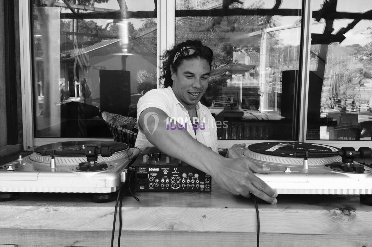 Un DJ mixant des vinyles sur une table de mixage en extérieur, avec un fond de fenêtre et de végétation.
