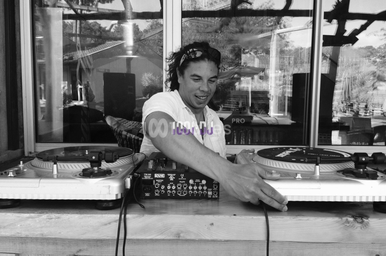 Un DJ mixant des vinyles sur une table de mixage en extérieur, avec un fond de fenêtre et de végétation.