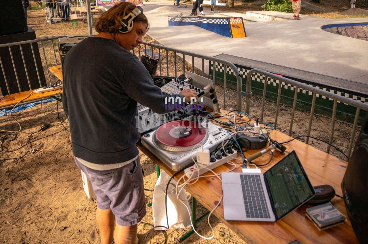 Un DJ mixe des vinyles sur une platine entourée d'équipements audio et d'un ordinateur en extérieur.