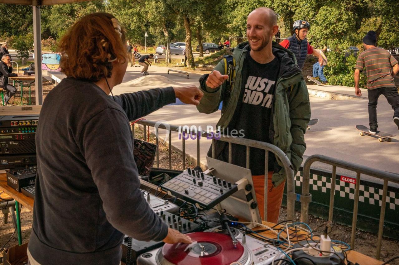 Un DJ serre le poing avec un homme souriant près d'une table de mixage, dans un skatepark animé en plein air.