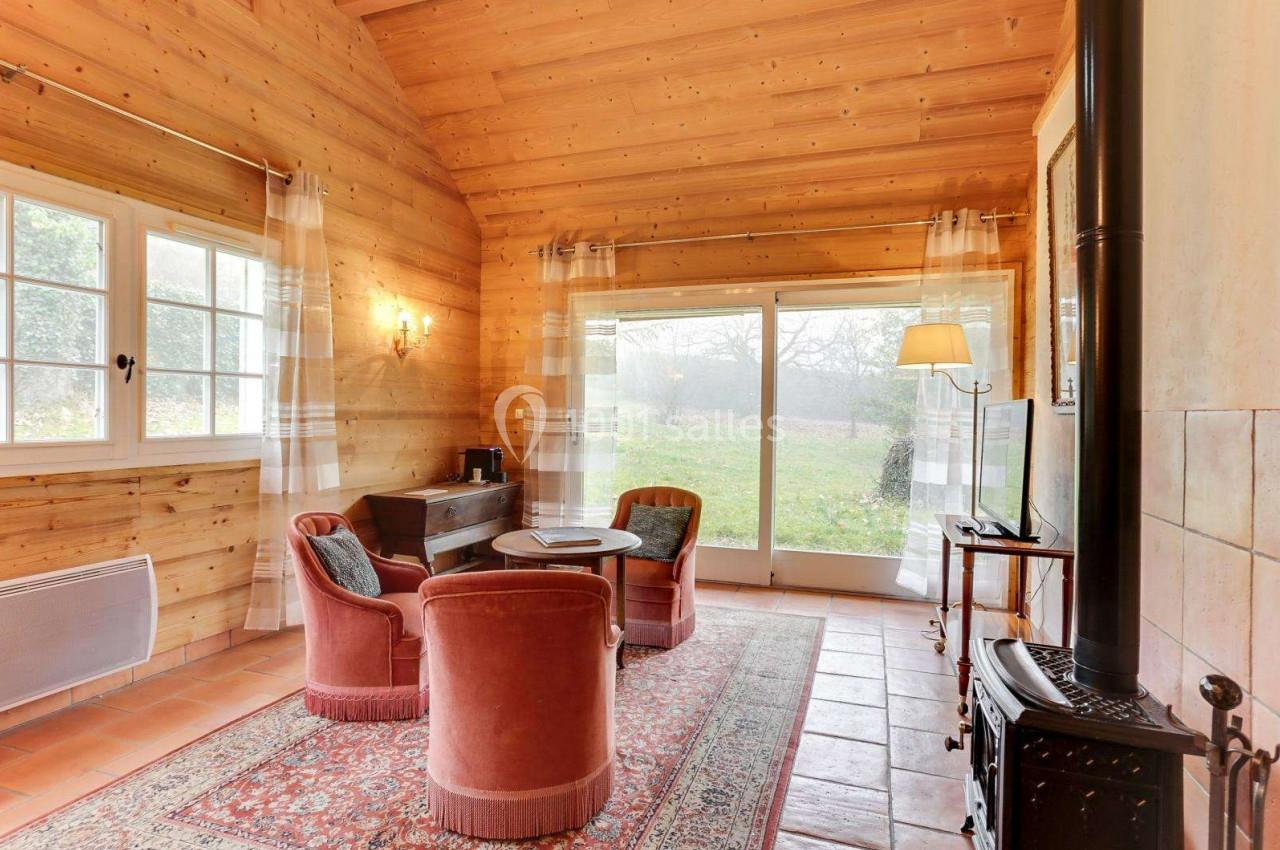 Salon chaleureux avec murs en bois, poêle à bois, fauteuils autour d'une table et vue sur un jardin par une baie vitrée.