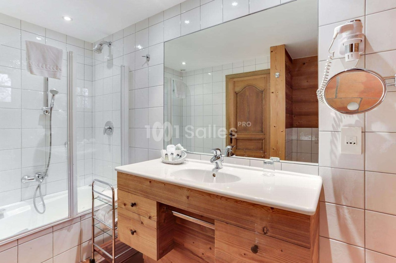 Salle de bain lumineuse avec baignoire, lavabo sur meuble en bois, miroir, sèche-cheveux et carrelage blanc.