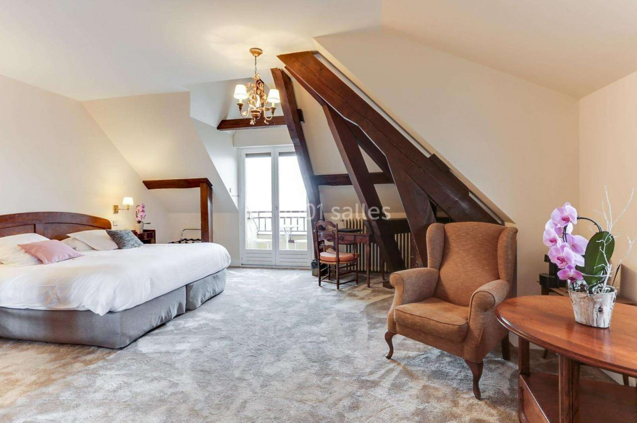 Chambre mansardée lumineuse avec lit double, fauteuil, table, orchidée et poutres apparentes, donnant sur un balcon.