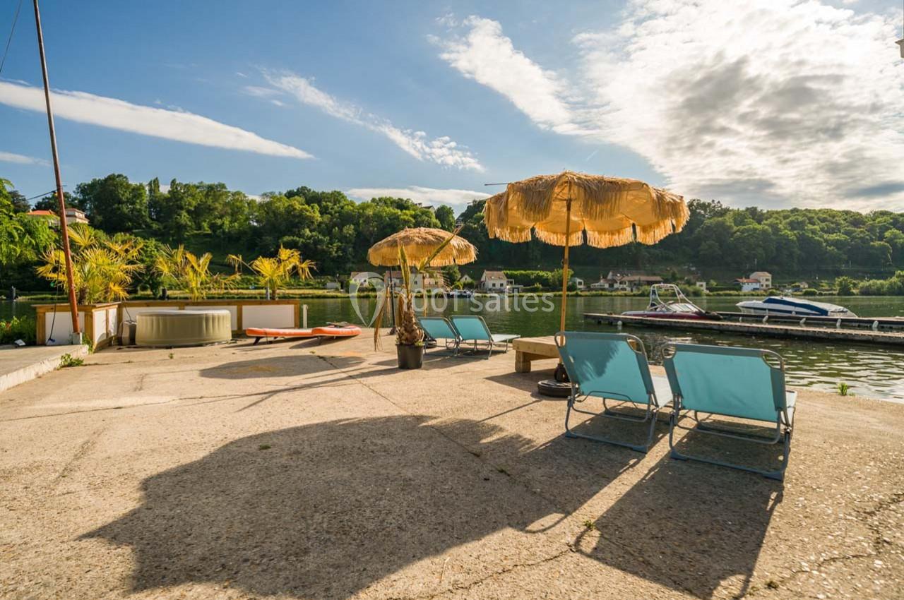 Chaises longues et parasols en bord de rivière, avec un quai, des bateaux et une végétation verdoyante en arrière-plan.