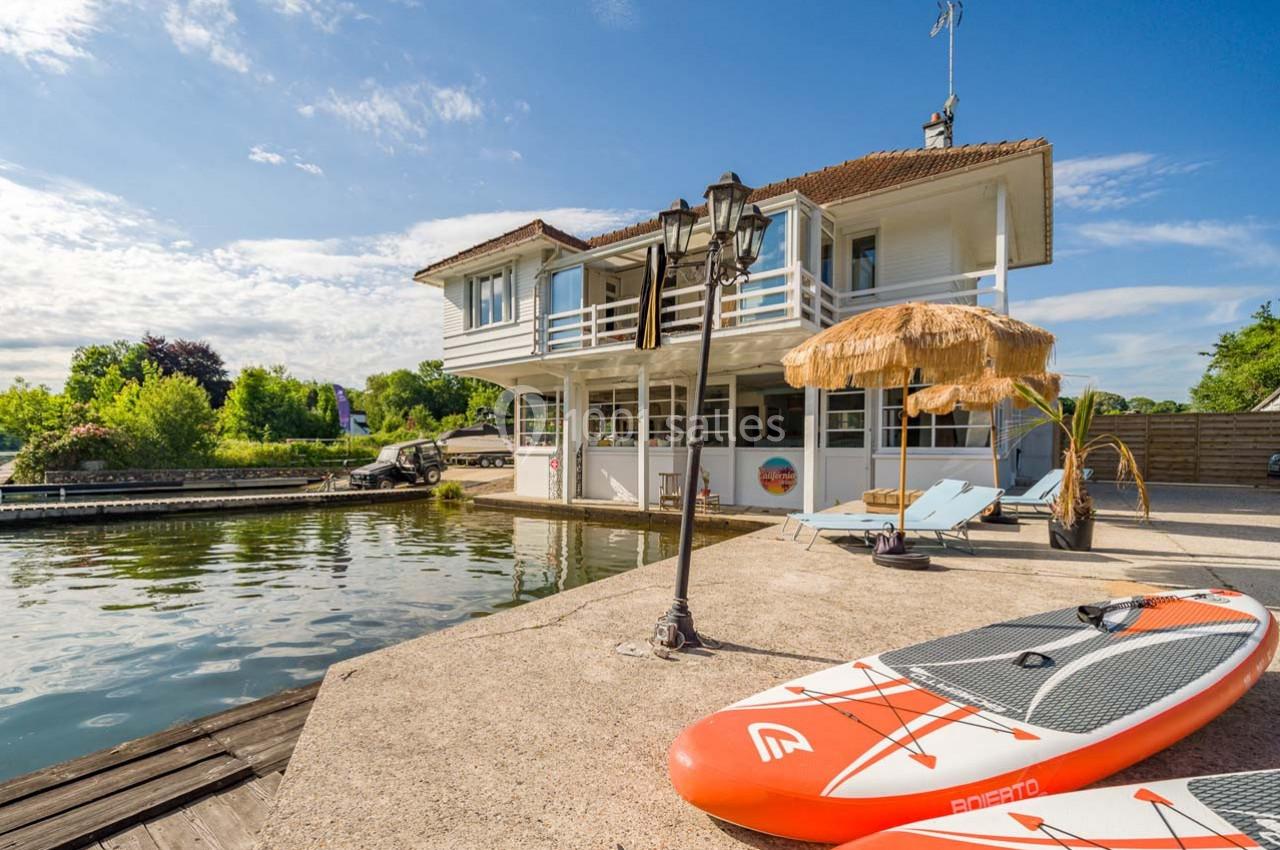 Maison au bord de l'eau avec terrasse, transats, parasol en paille et planche de paddle sur un quai ensoleillé.