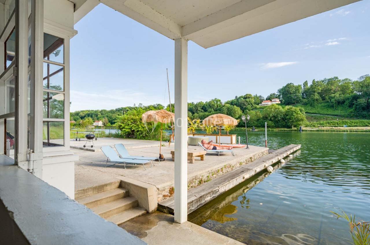Terrasse au bord d'une rivière avec transats, parasols en paille et vue sur un paysage verdoyant.
