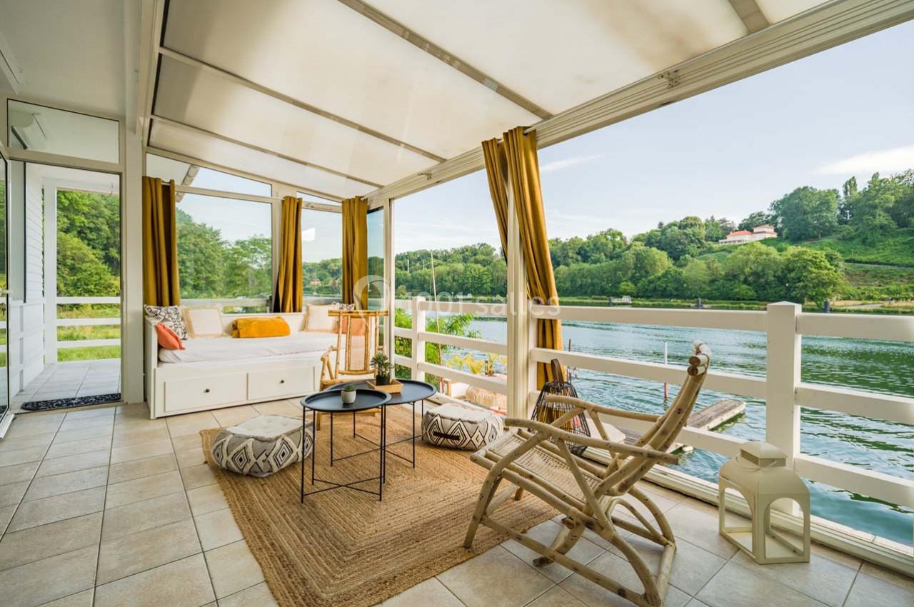 Véranda lumineuse avec vue sur une rivière, aménagée avec des fauteuils en rotin, un tapis et des coussins colorés.