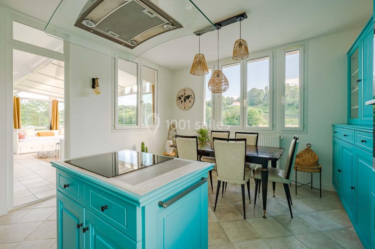 Cuisine lumineuse avec meubles turquoise, table à manger en bois foncé, grandes fenêtres et vue sur un paysage verdoyant.