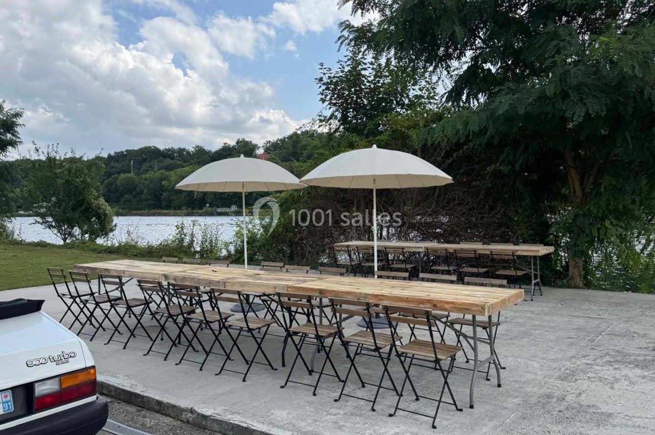 Tables et chaises en bois disposées en extérieur près d'une rivière, avec parasols et végétation en arrière-plan.