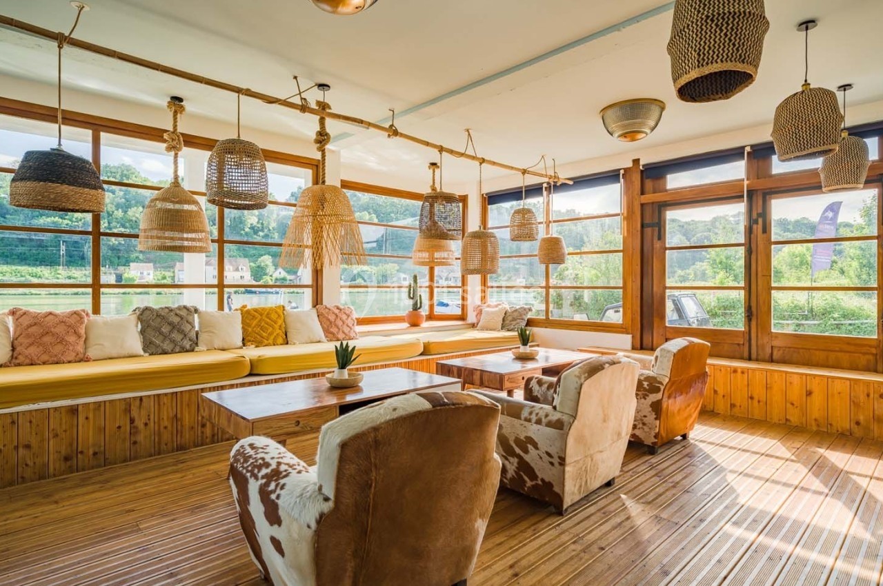 Salle lumineuse avec de grandes fenêtres, fauteuils en cuir, banquette jaune et suspensions en osier.