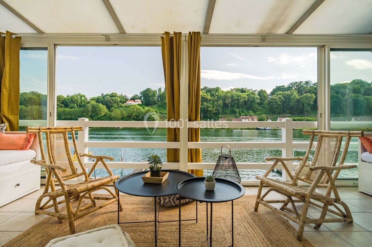 Salon lumineux avec mobilier en rotin, tables basses noires et vue sur un lac entouré de verdure à travers de grandes baies…