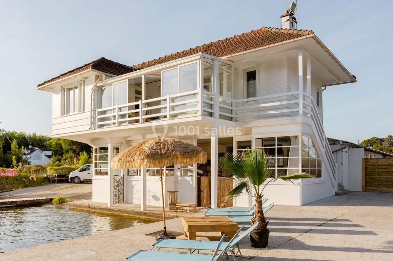 Maison blanche à étage avec terrasse, toit en tuiles, palmiers et chaises longues près d'un plan d'eau.