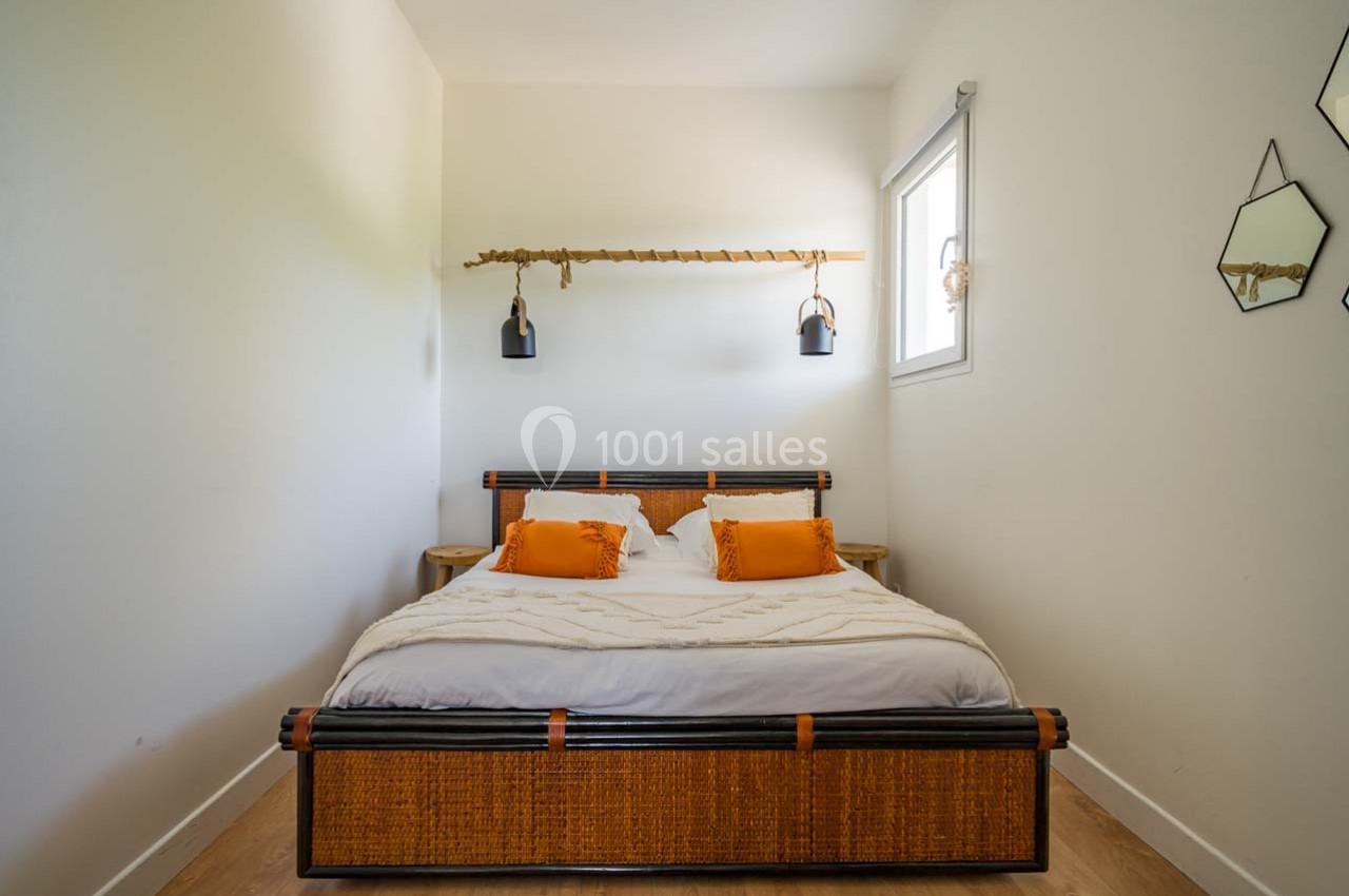 Lit double avec tête de lit en rotin, coussins orange, éclairage suspendu et mur décoré de miroirs géométriques.