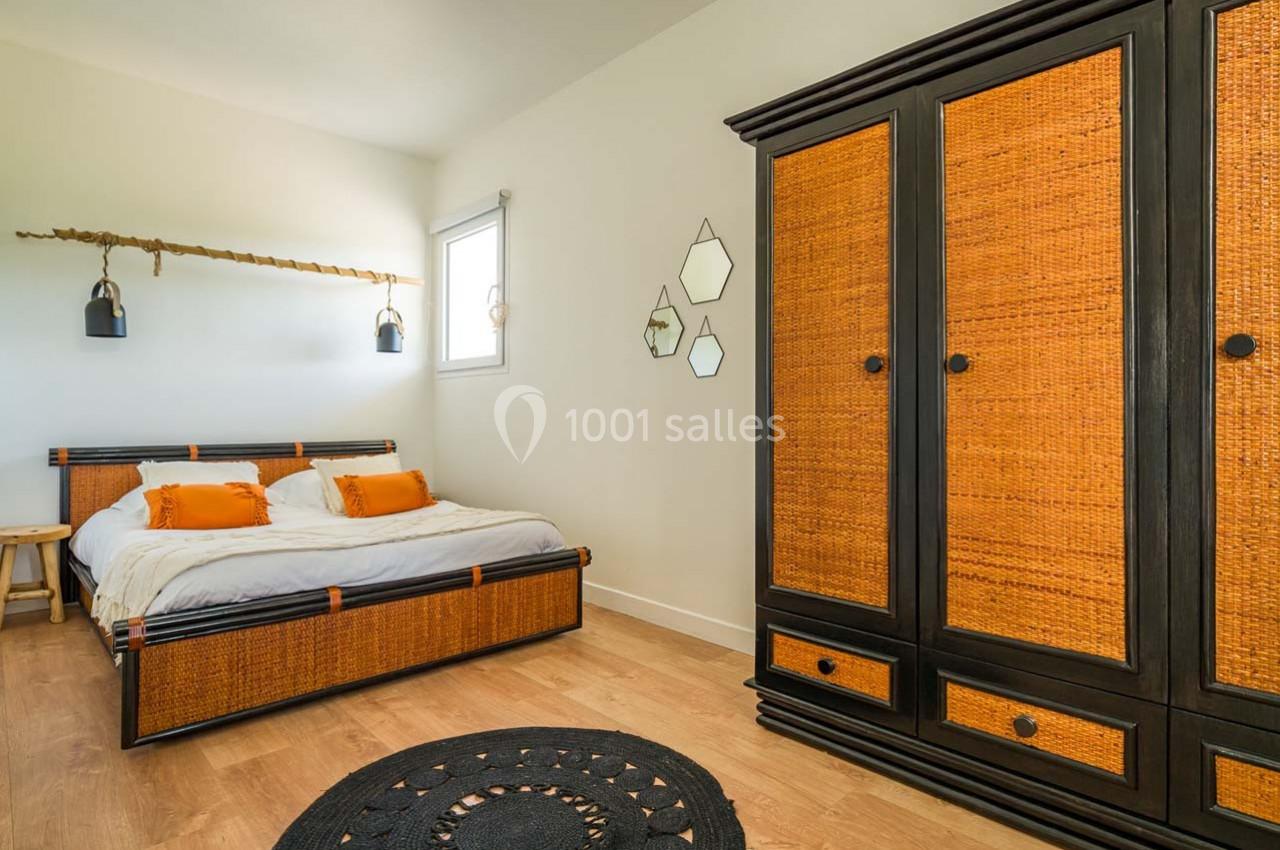Chambre avec lit double et armoire en rotin, murs clairs, décor minimaliste et tapis rond noir au sol.