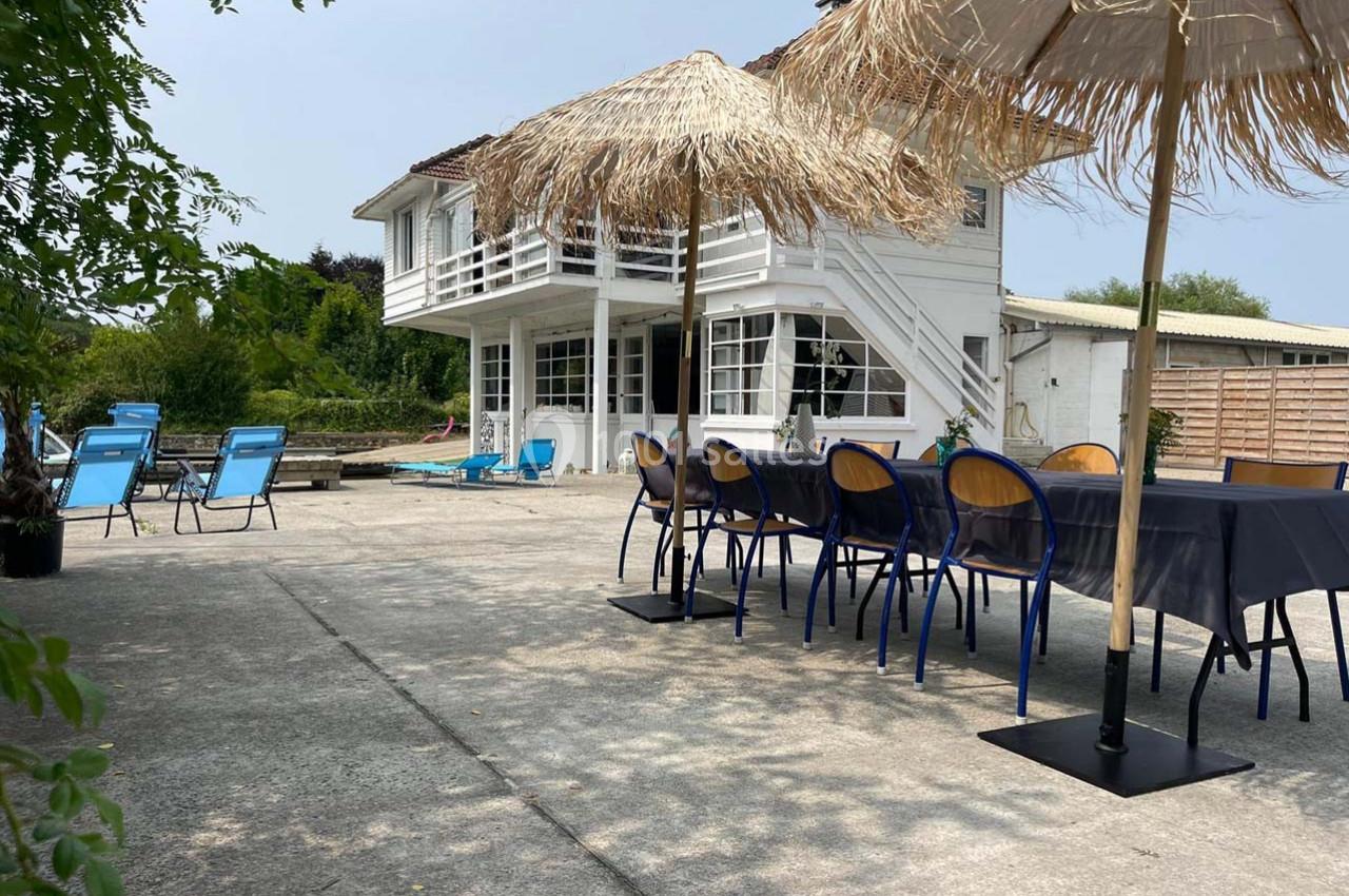 Terrasse ensoleillée avec table, chaises bleues et parasols en paille, devant une maison blanche à deux étages.