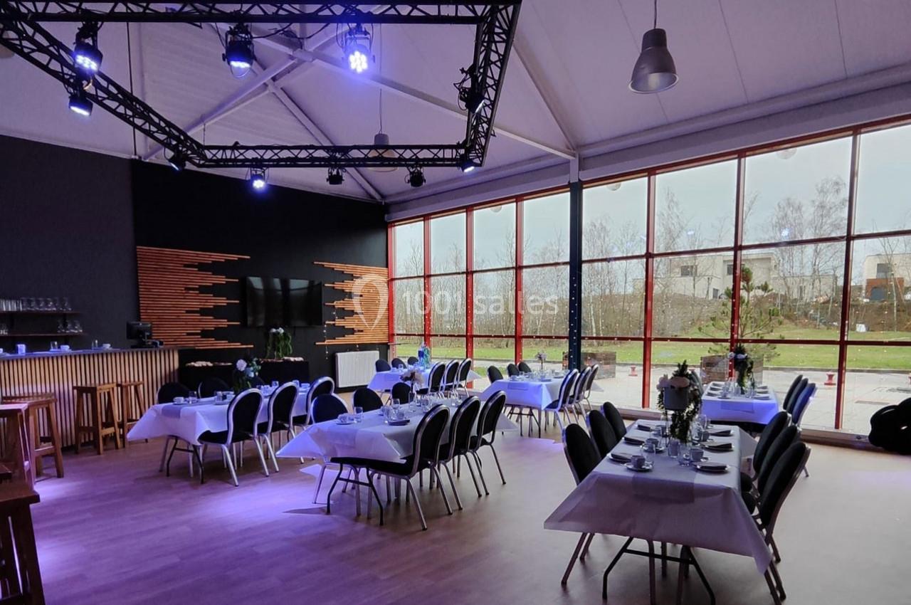 Salle lumineuse aménagée pour un événement, avec des tables dressées, des chaises noires et une grande baie vitrée.