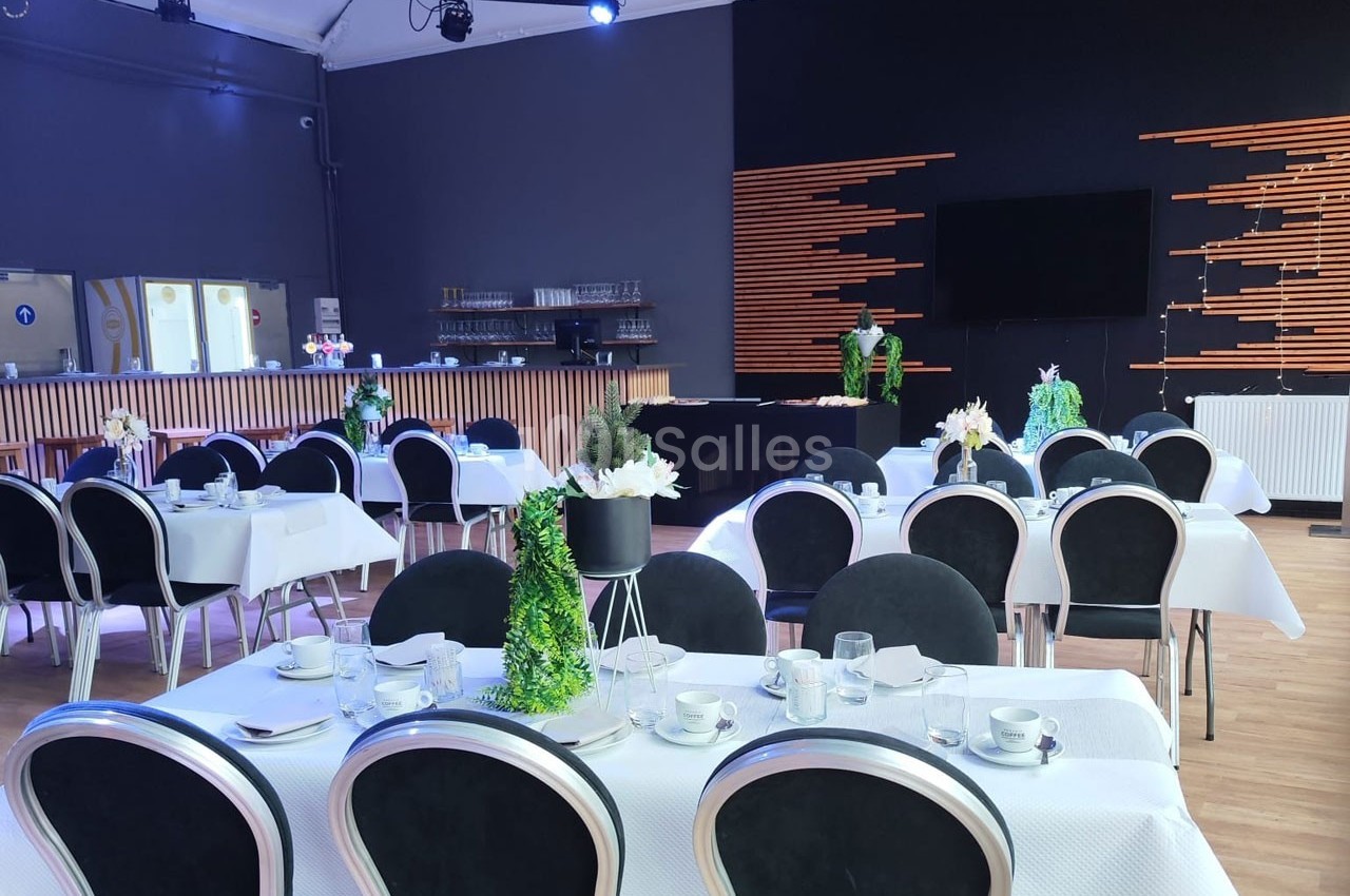 Salle de réception avec tables dressées, chaises noires, décorations végétales et un comptoir en arrière-plan.