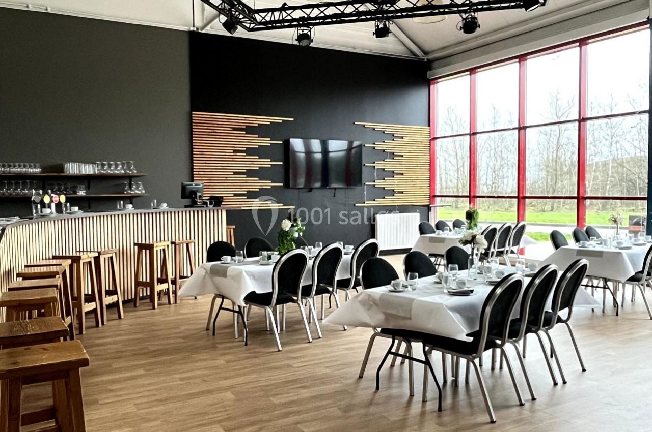 Salle de restaurant lumineuse avec tables dressées, chaises noires, bar en bois et grande baie vitrée donnant sur l…