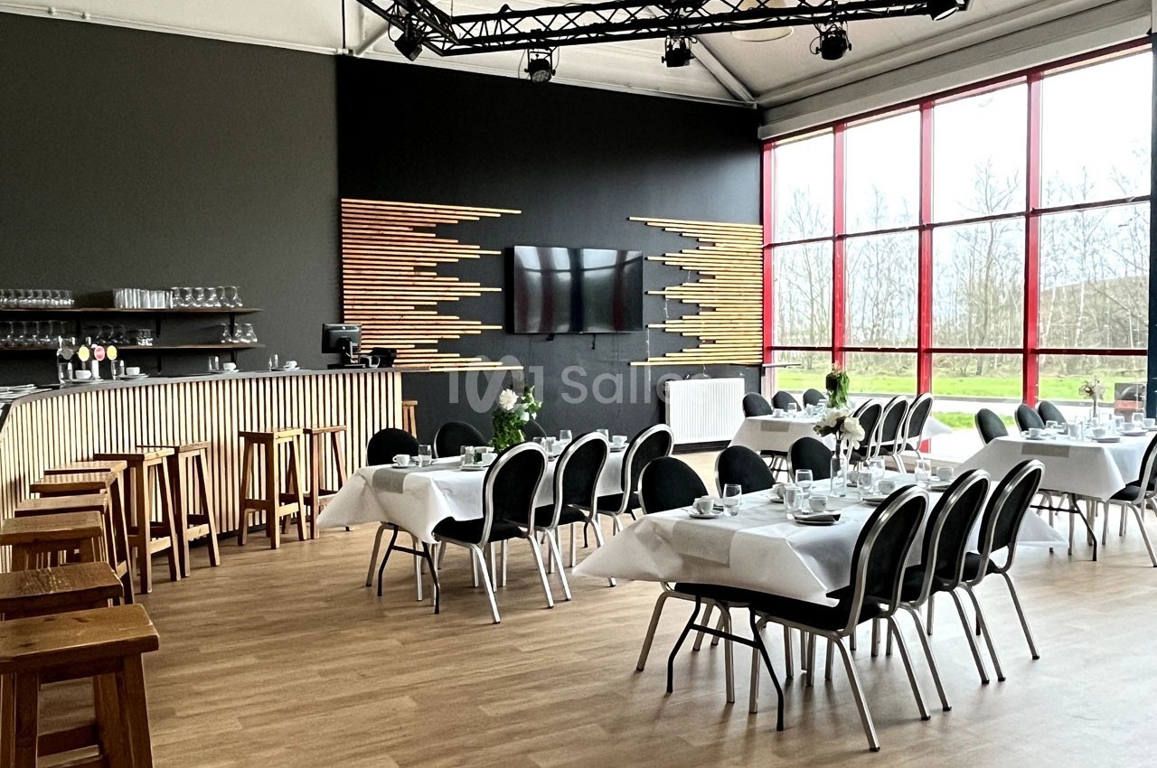 Salle de restaurant lumineuse avec tables dressées, chaises noires, bar en bois et grande baie vitrée donnant sur l…