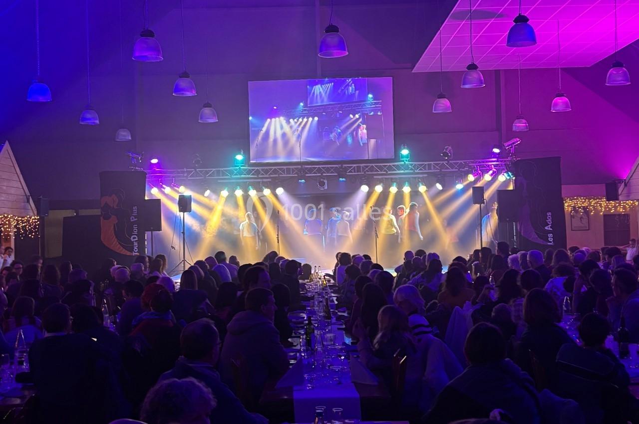 Salle comble avec des tables alignées, éclairage coloré et scène illuminée au fond diffusant un spectacle.