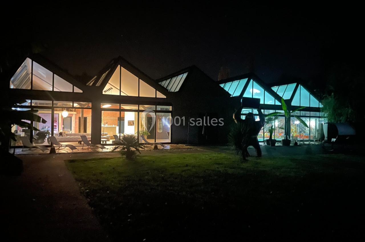 Façade vitrée d'une maison moderne éclairée de nuit, entourée de végétation et d'une pelouse sombre.