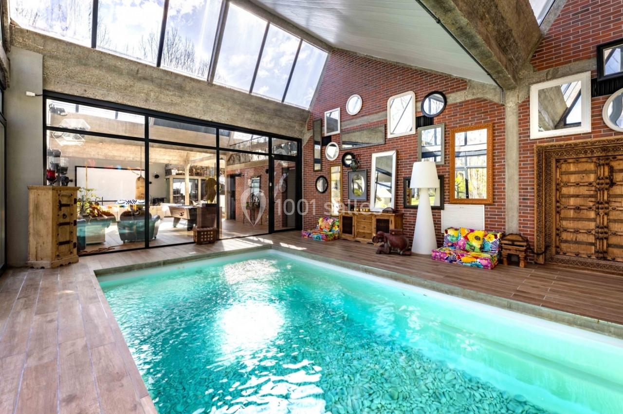 Piscine intérieure lumineuse avec sol en bois, murs en briques, grandes baies vitrées et décoration éclectique.