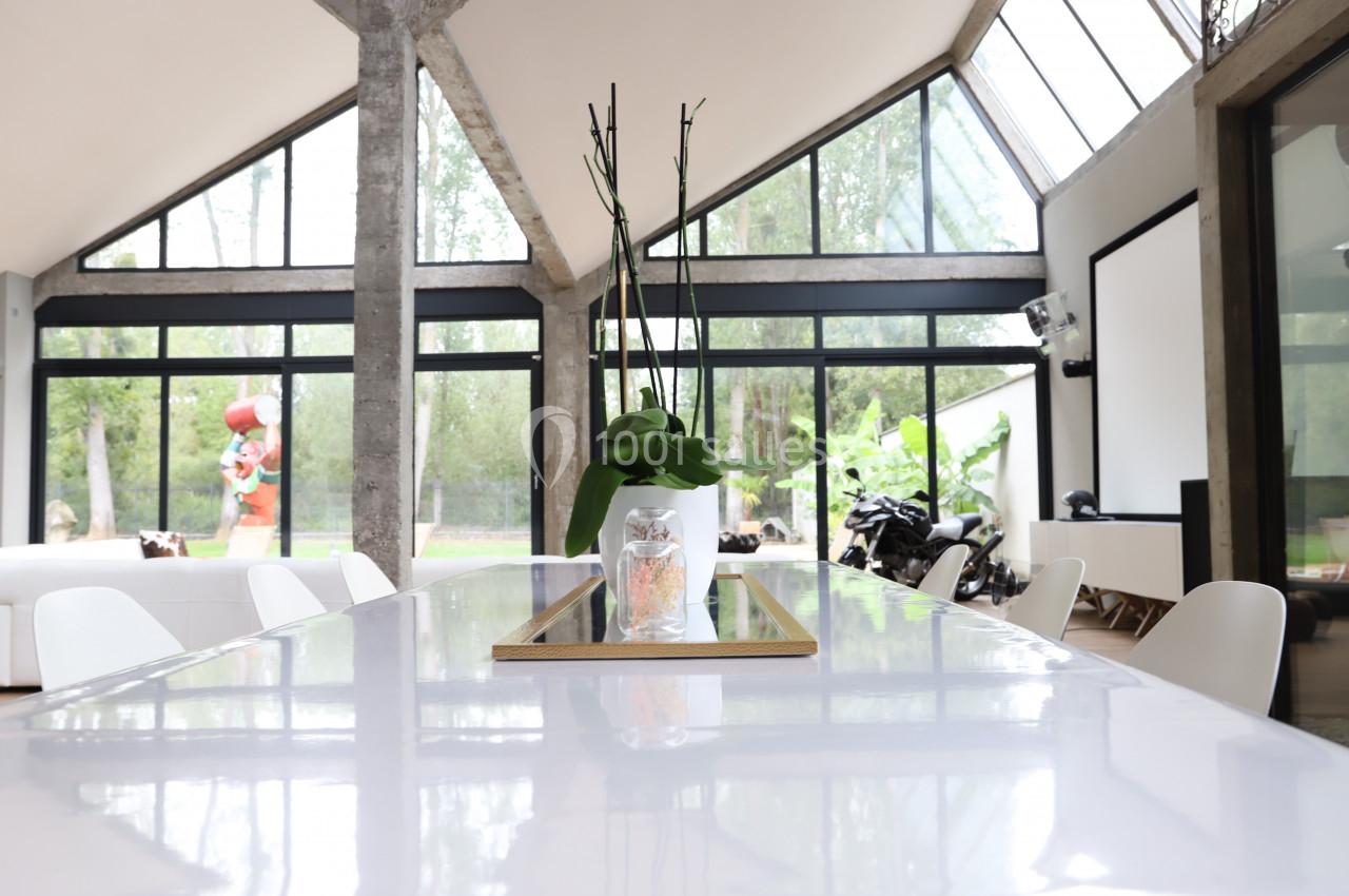 Table blanche avec un vase contenant une plante, dans une pièce lumineuse aux grandes baies vitrées donnant sur un jardin.