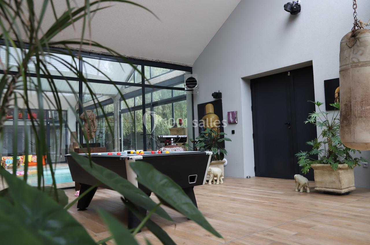 Salle lumineuse avec table de billard, plantes, objets décoratifs et vue sur une piscine intérieure à travers des baies…