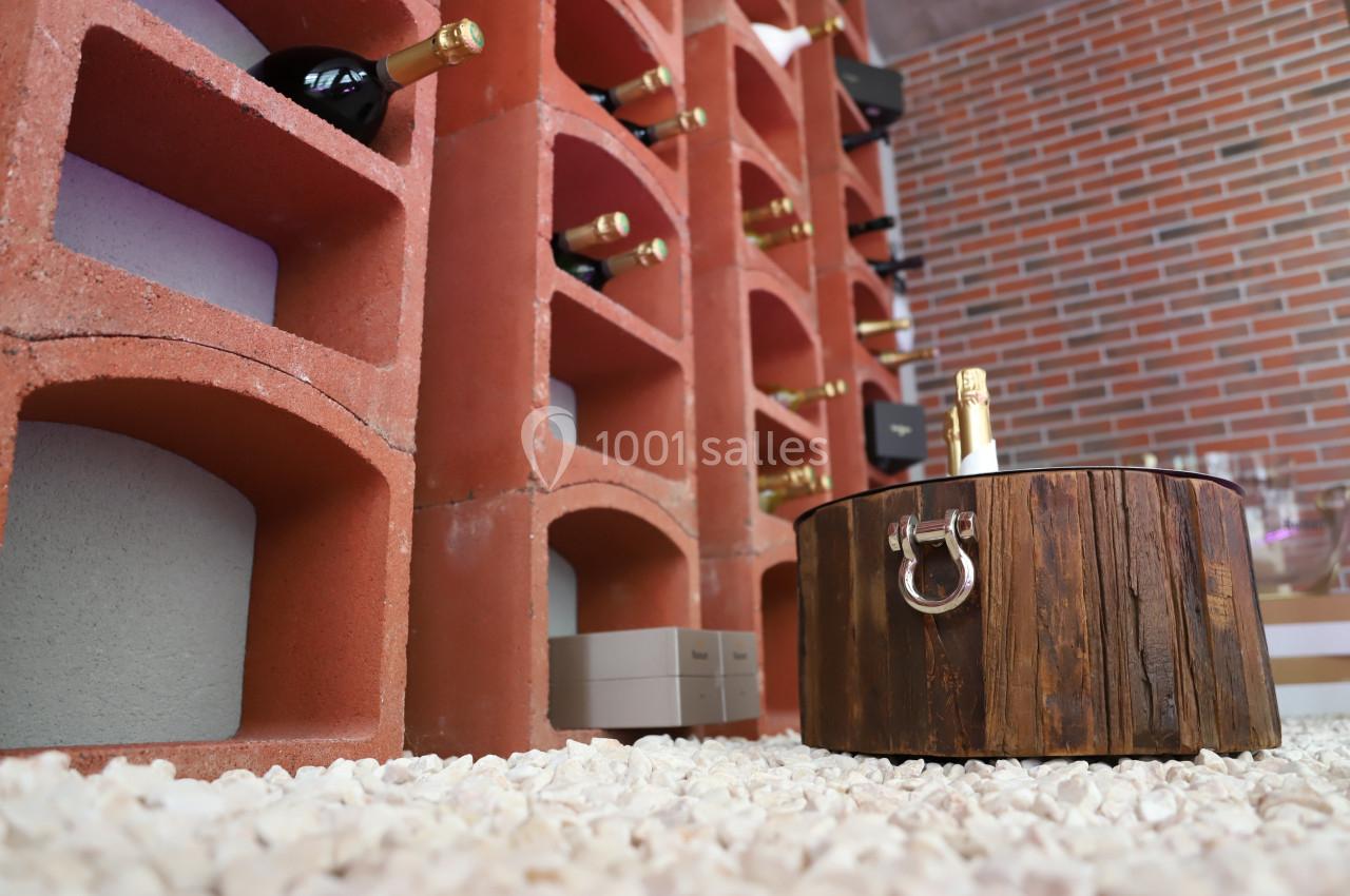 Casiers en briques rouges contenant des bouteilles de vin, avec un seau en bois au premier plan sur un sol en gravier.