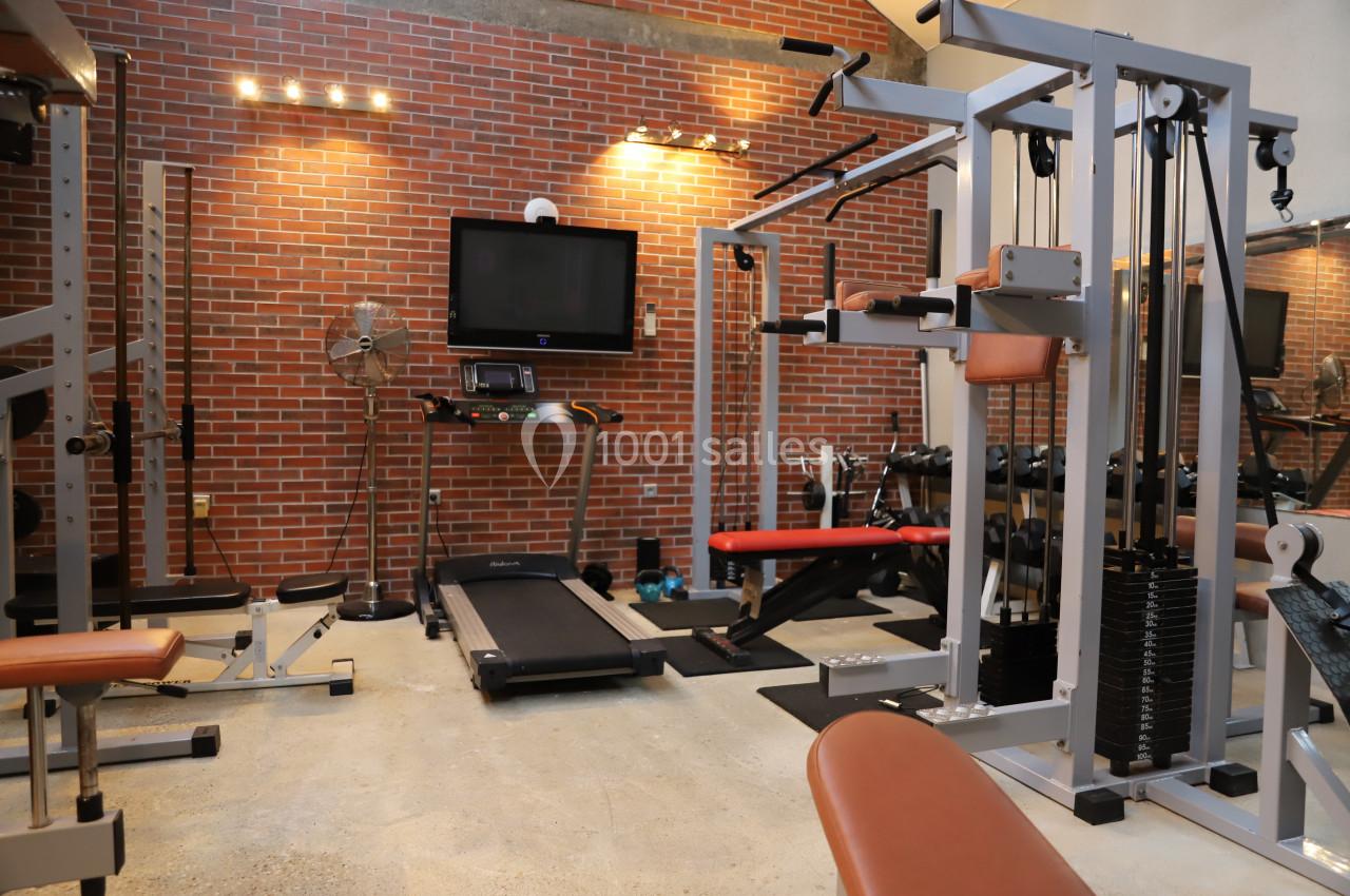 Salle de sport équipée de machines de musculation, tapis de course et bancs, avec mur en briques en arrière-plan.