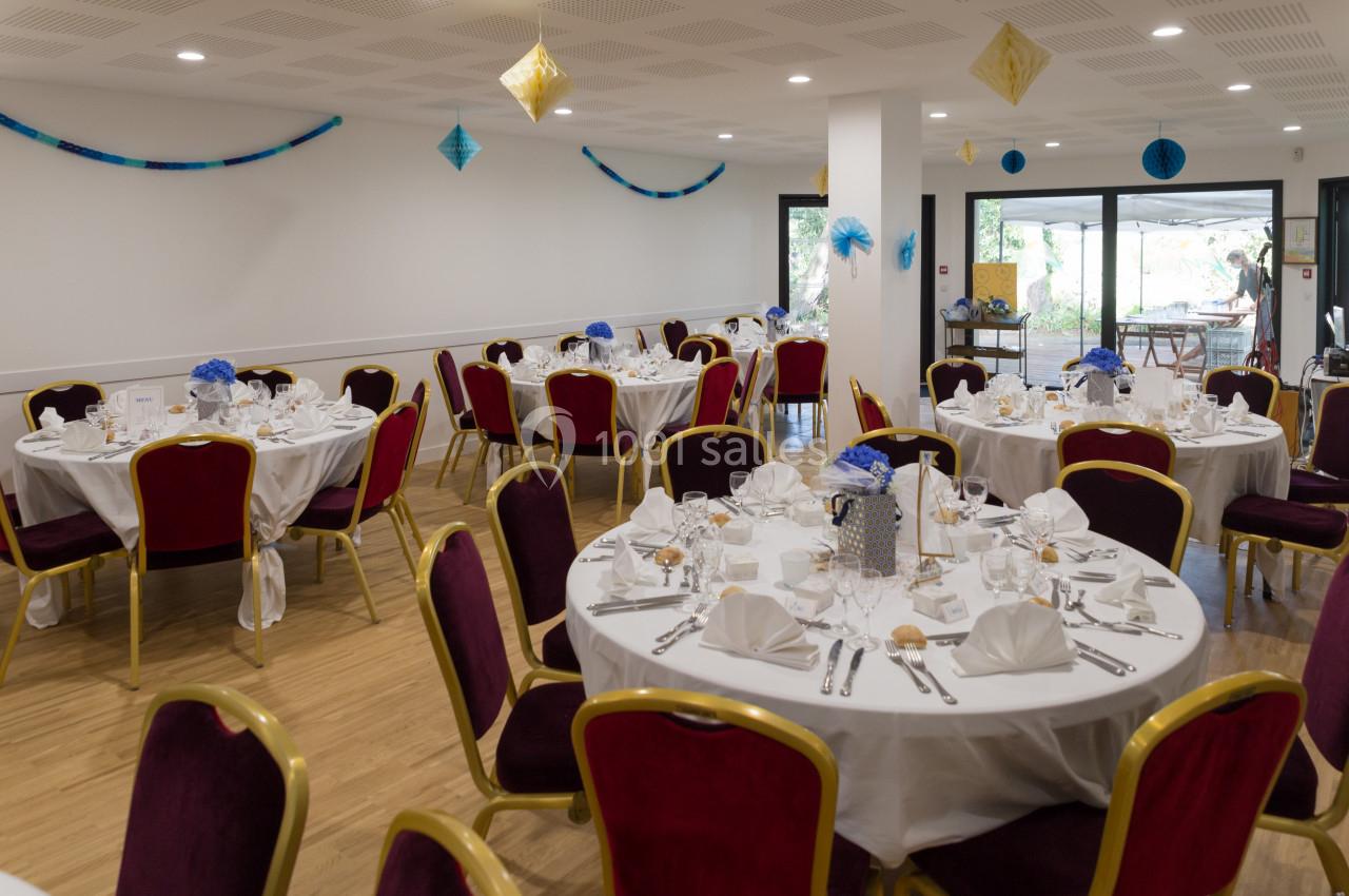 Salle de réception décorée avec des tables rondes dressées, nappes blanches et chaises rouges, prête pour un événement.