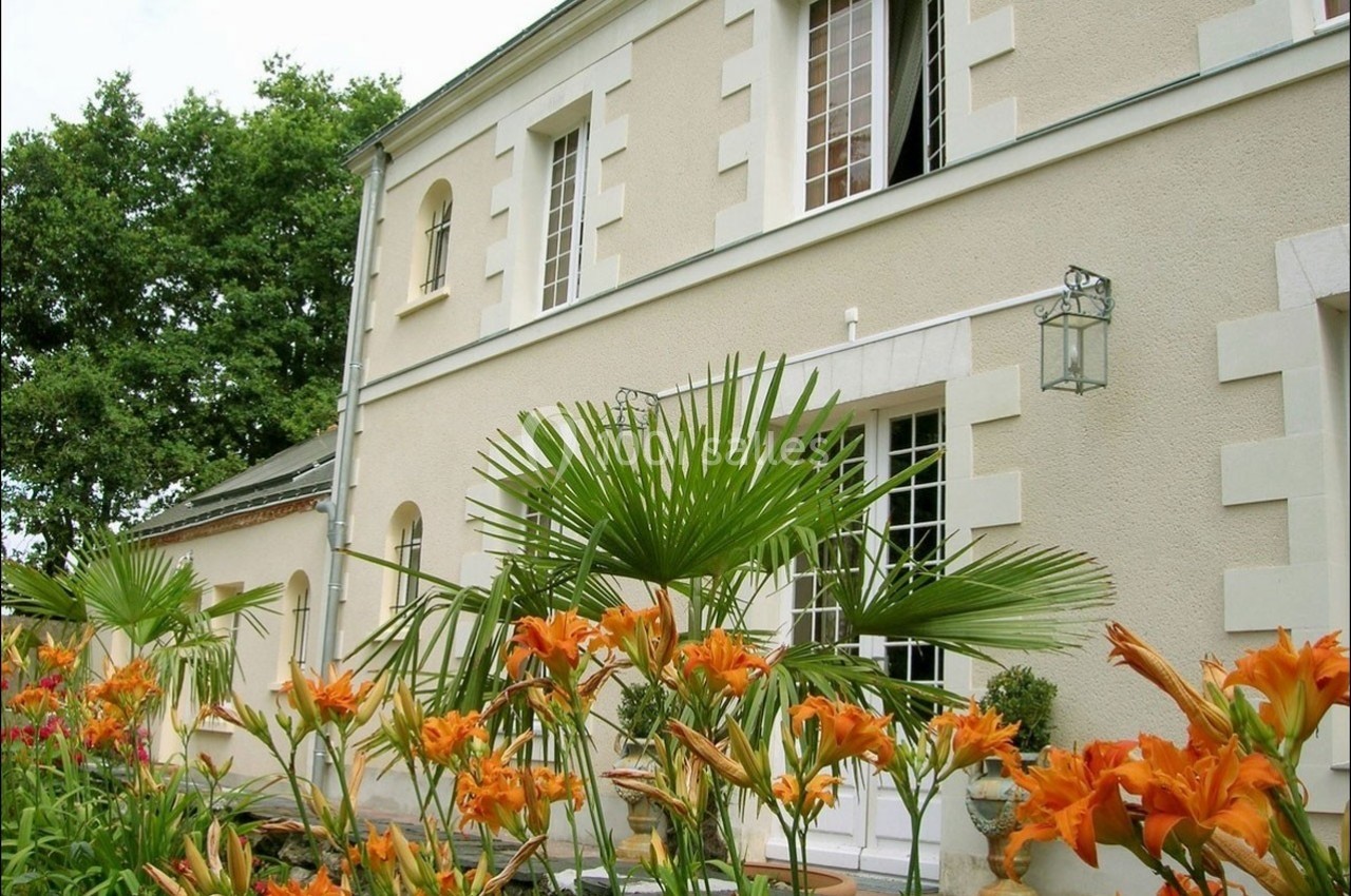 Façade d'une maison beige avec des fenêtres blanches, entourée de fleurs orange et de palmiers sous un ciel clair.