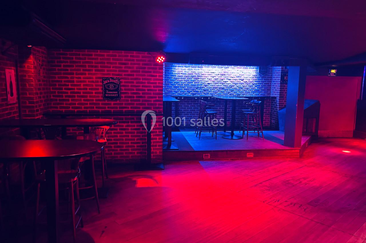 Salle de bar avec murs en briques, éclairage rouge et bleu, tables hautes et une petite scène surélevée.