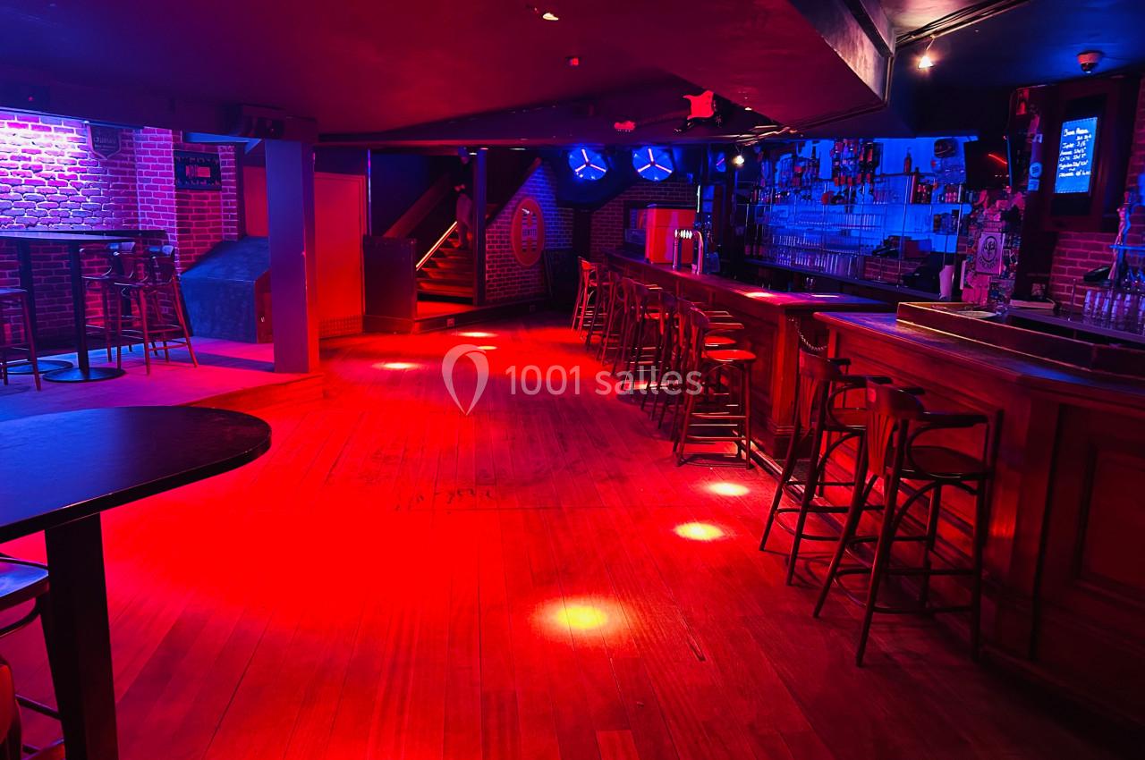 Salle de bar avec éclairage rouge tamisé, comptoir en bois, tabourets alignés et tables hautes dispersées.