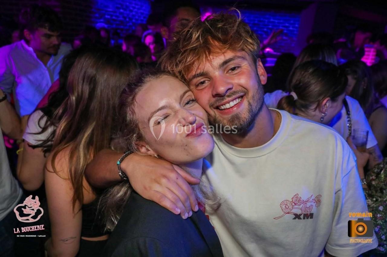 Un homme et une femme souriants posent ensemble dans une ambiance festive avec des personnes en arrière-plan.
