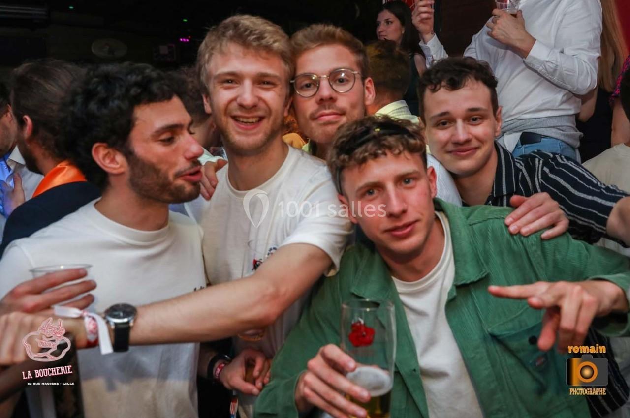 Cinq jeunes hommes souriants posent ensemble dans un environnement festif, certains tenant des verres à la main.