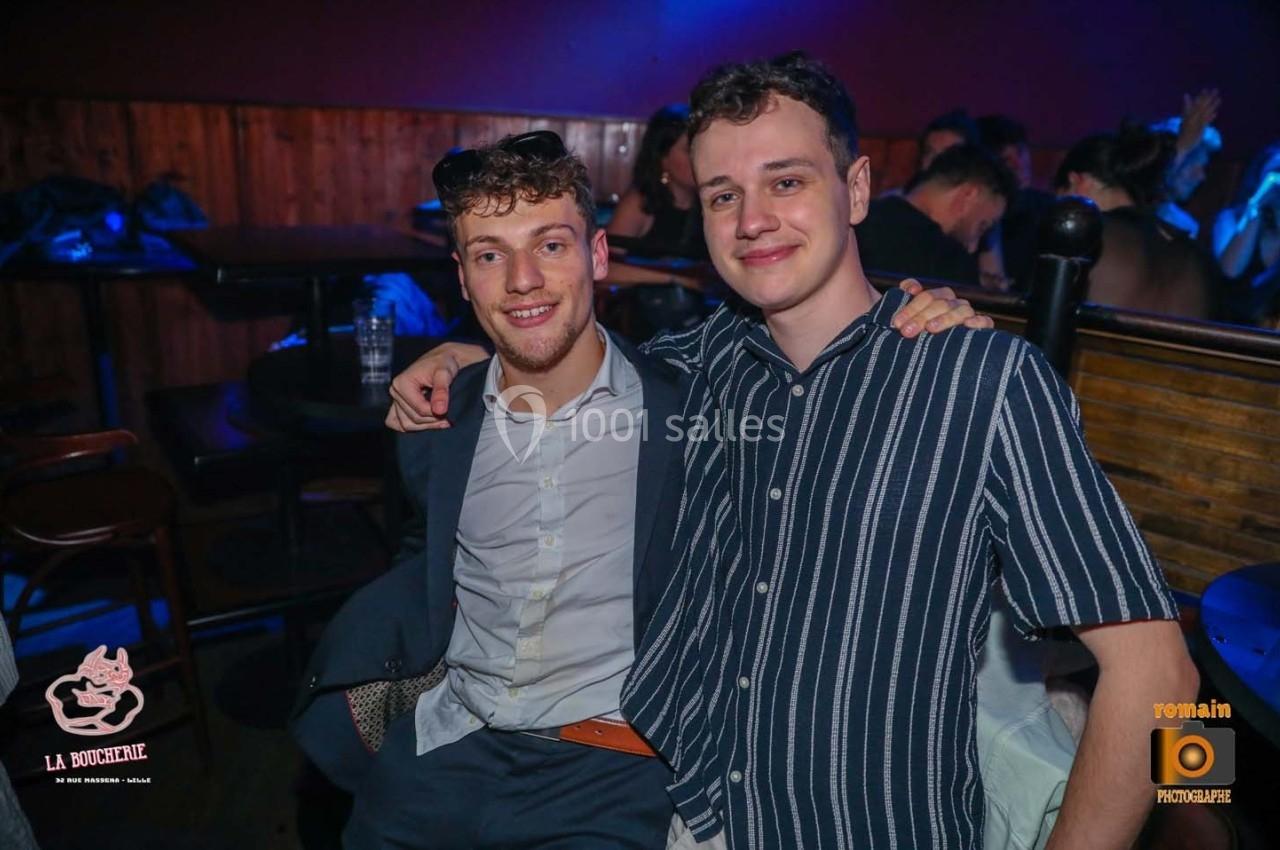 Deux hommes souriants posent côte à côte dans un bar à l'ambiance tamisée.