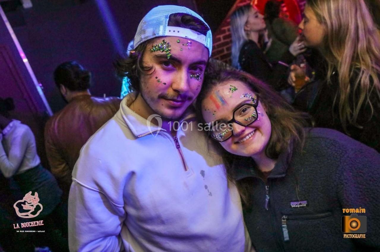 Deux personnes souriantes avec des maquillages colorés et pailletés dans un environnement festif.