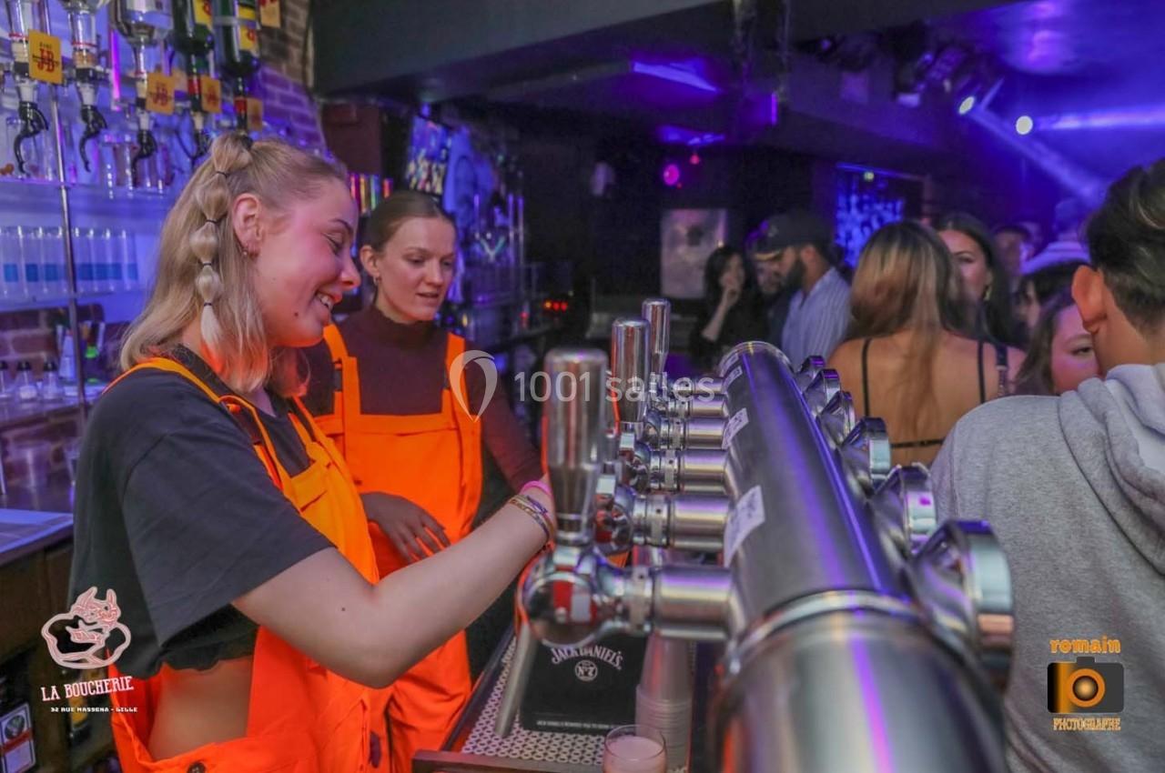 Deux serveuses en tabliers orange servent des boissons derrière un bar dans une ambiance de soirée animée.