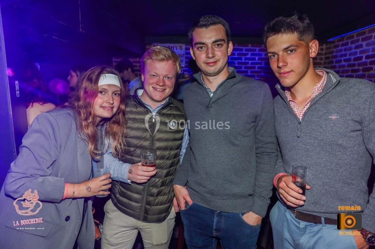 Quatre jeunes adultes posent ensemble dans un bar, tenant des verres, avec un éclairage tamisé et des logos visibles en…