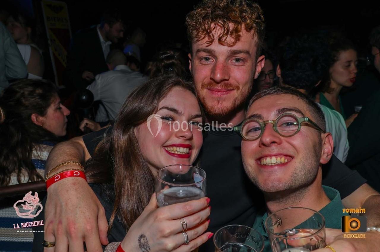 Trois personnes souriantes tenant des verres dans un environnement festif et animé.