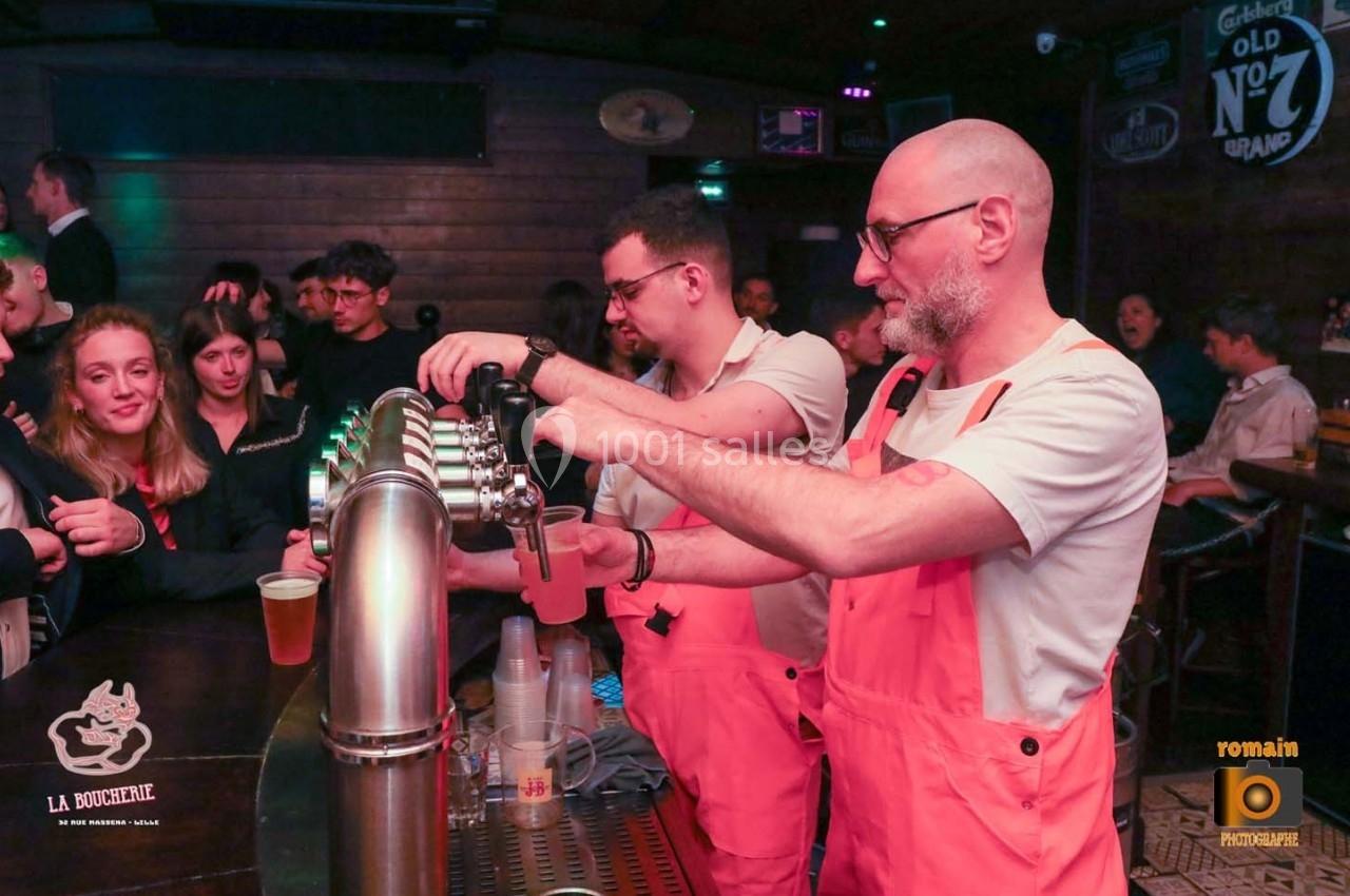 Deux barmans en tabliers roses servent des boissons à un comptoir dans un bar animé.