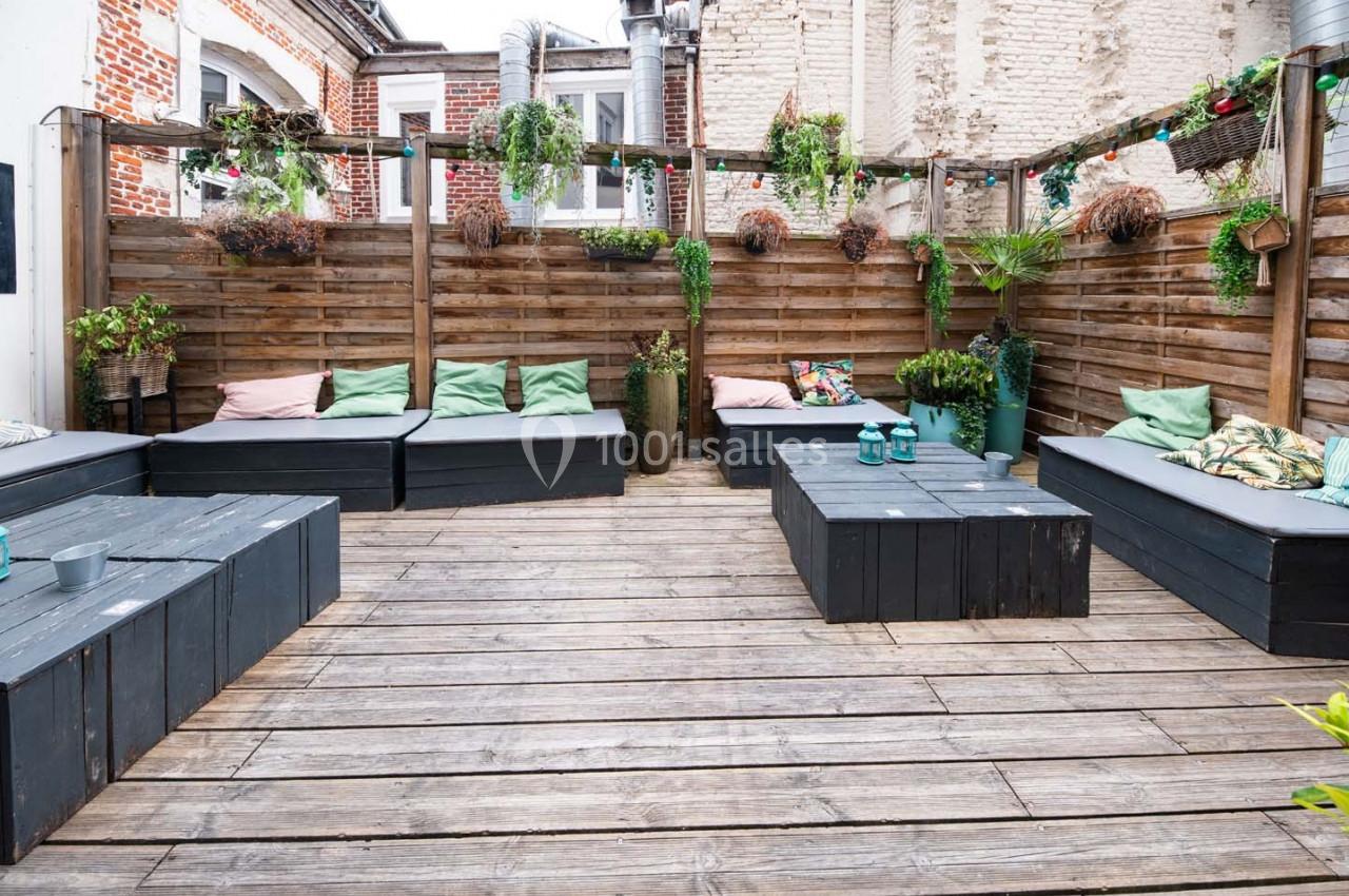 Terrasse en bois avec banquettes noires, coussins colorés et plantes suspendues dans un espace extérieur clos.