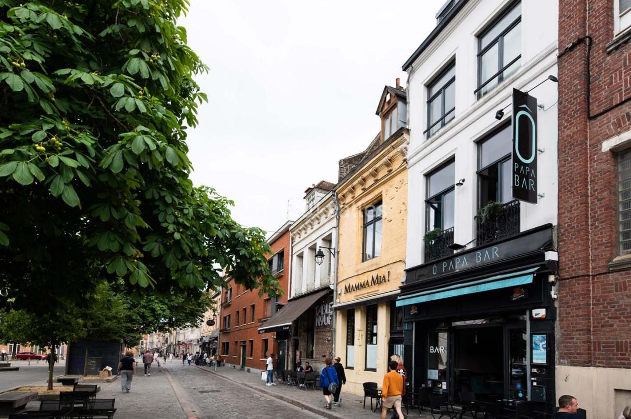 Rue pavée bordée de bâtiments, dont un bar et des restaurants, avec des passants et un arbre à gauche.