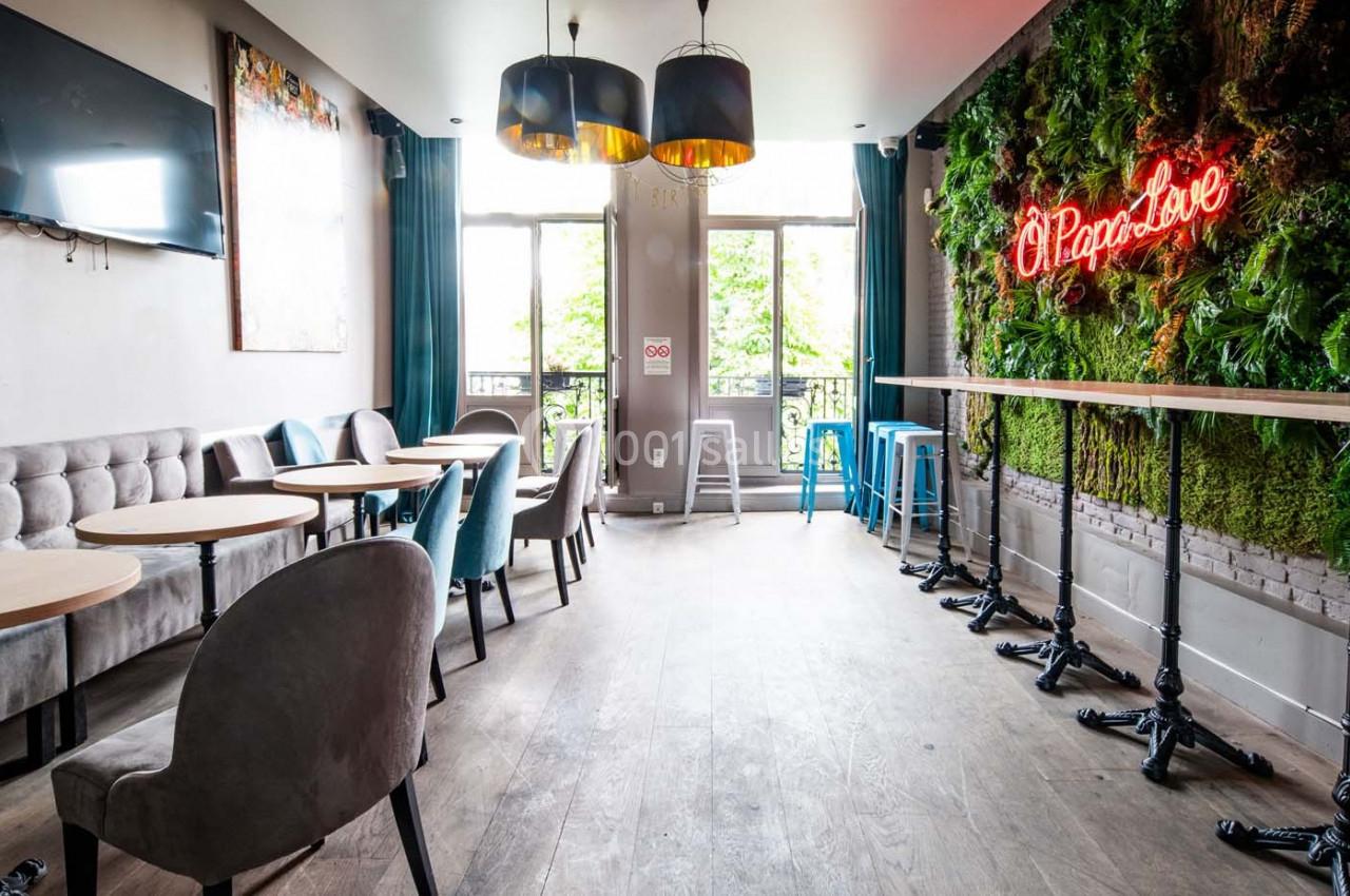 Salle lumineuse avec tables, chaises, mur végétal, enseigne néon et grandes fenêtres donnant sur l'extérieur.